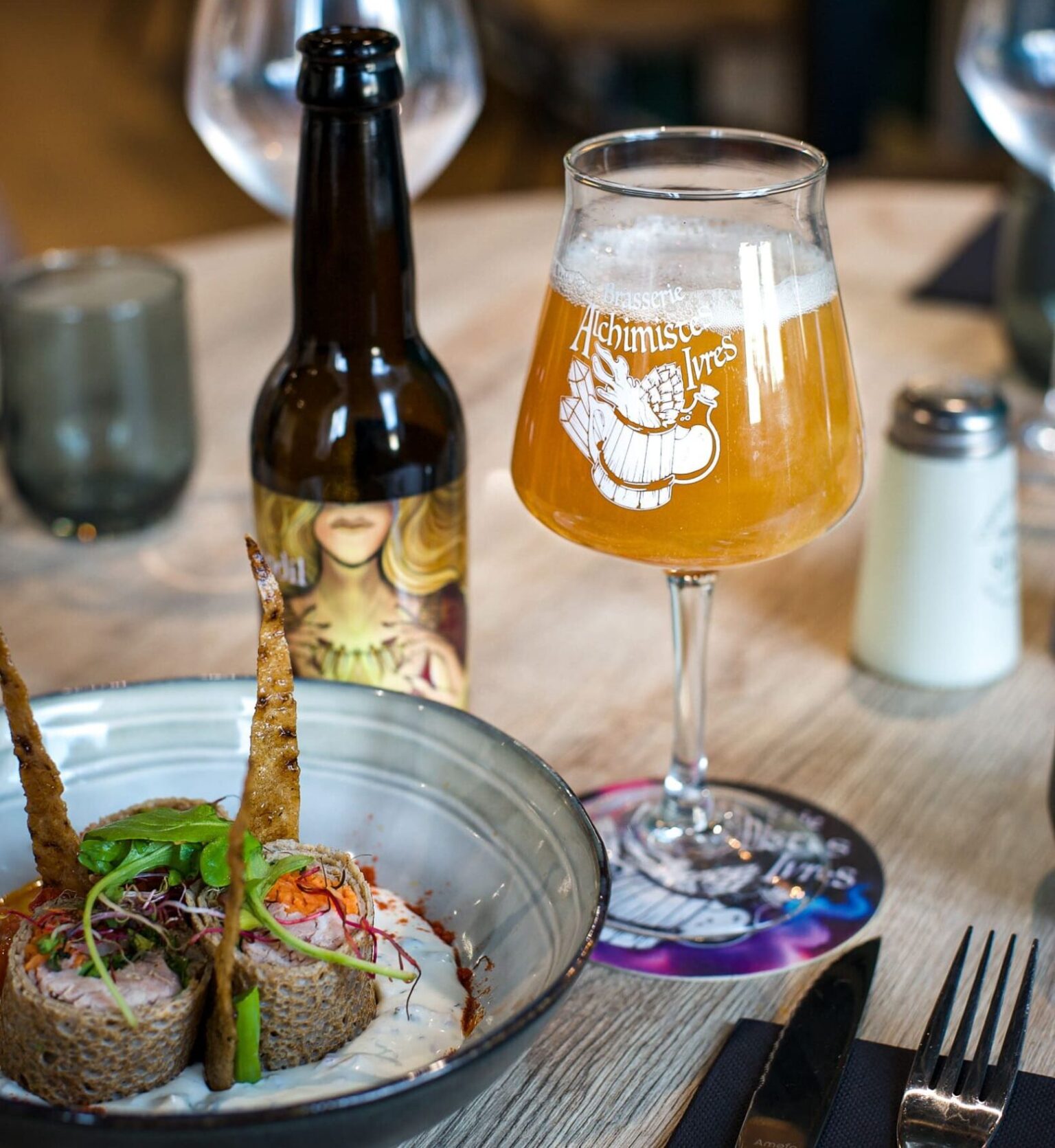 Verre et bouteille de bière artisanale Les Alchimistes Ivres servis à table, accompagnés d’un plat gastronomique pour un accord mets et bière.